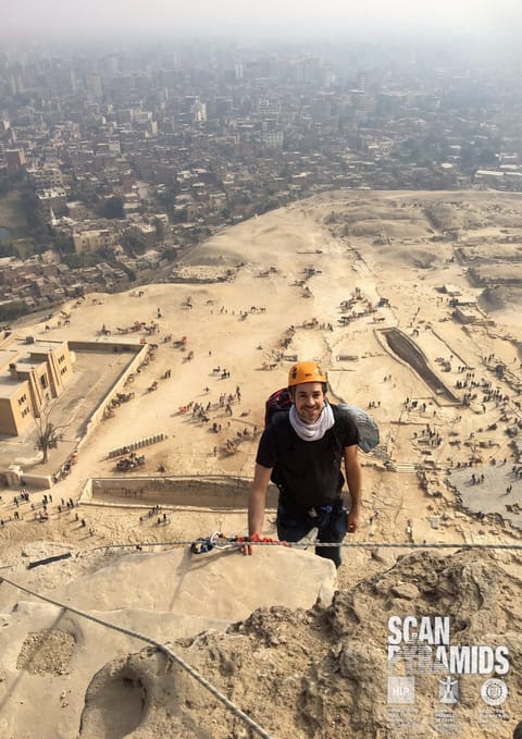 Benoit Marini, casque orange et corde, sur le flanc d'une pyramide, Le Caire en contrebas