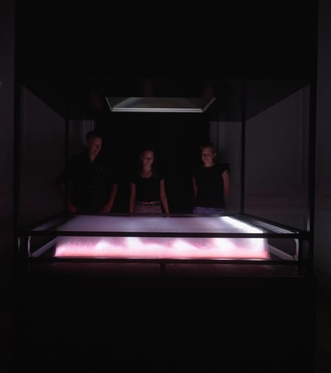 Trois visiteurs dans la pénombre regardent Turbulence, une nappe de lumière rose posée devant eux